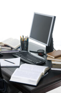 Desk with open books, pens, notepad, monitor and keyboard.
